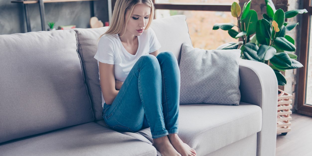 Upset woman has first symptoms of pms. She is sitting on a sofa and touching her stomach. She is going to cry.