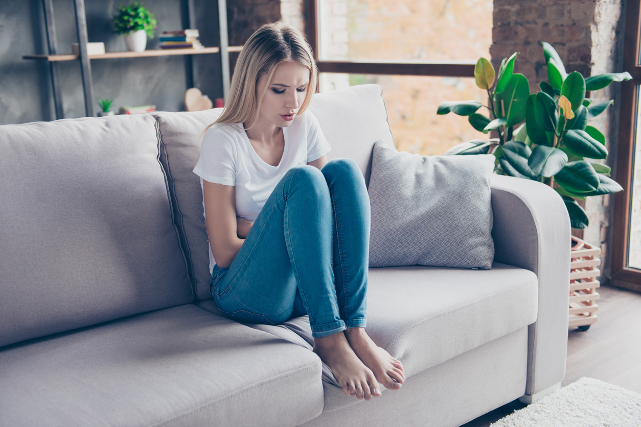 Upset woman has first symptoms of pms. She is sitting on a sofa and touching her stomach. She is going to cry.